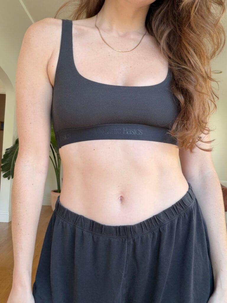 10 Best Sustainable Underwear Brands (2025 Review) A woman wearing a black sports bra and black pants stands indoors, partially facing the camera.