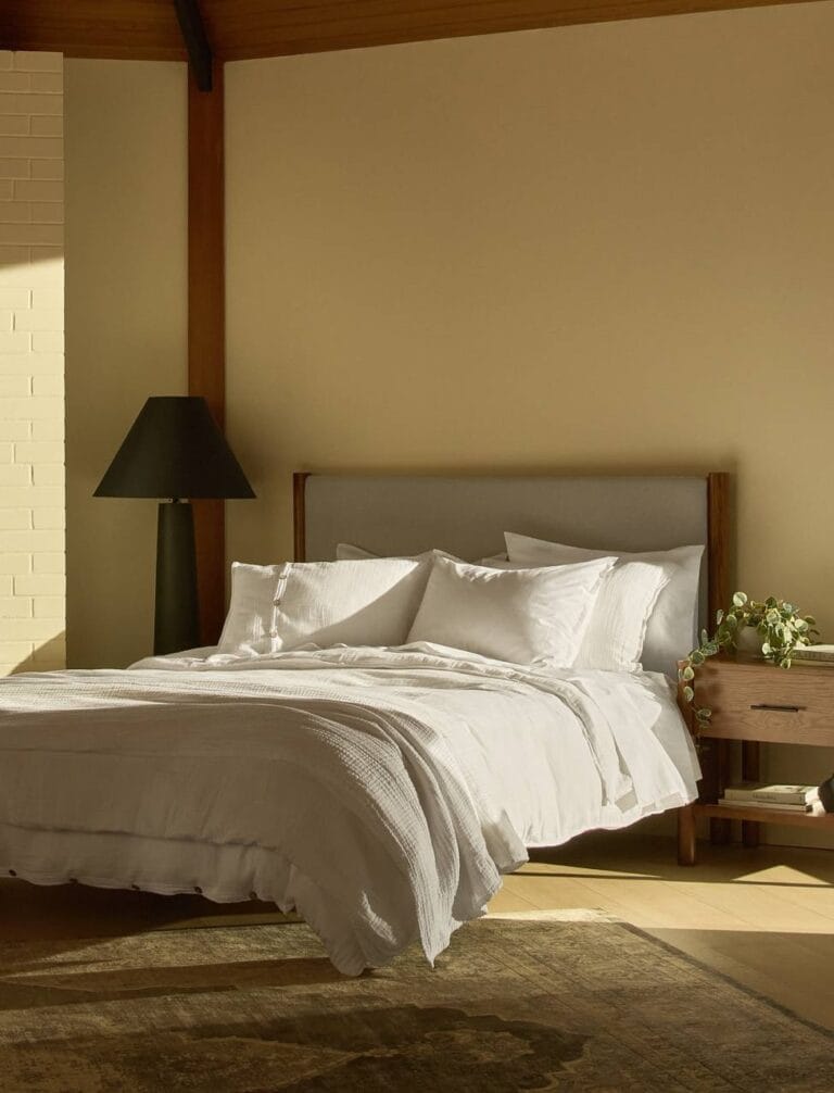 A neatly made bed with white linens and multiple pillows sits beside a wooden nightstand with a vase of flowers, under soft natural light in a minimalist bedroom.