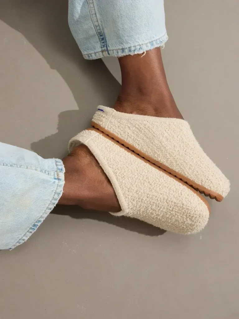 Person wearing light blue jeans and cozy, cream-colored slippers with textured fabric and rubber soles, resting feet on a neutral gray floor.
