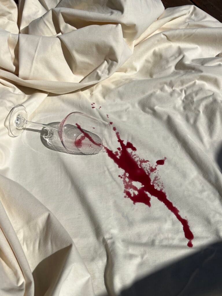 A tipped-over wine glass with red wine spilled onto a white sheet, creating a noticeable stain.