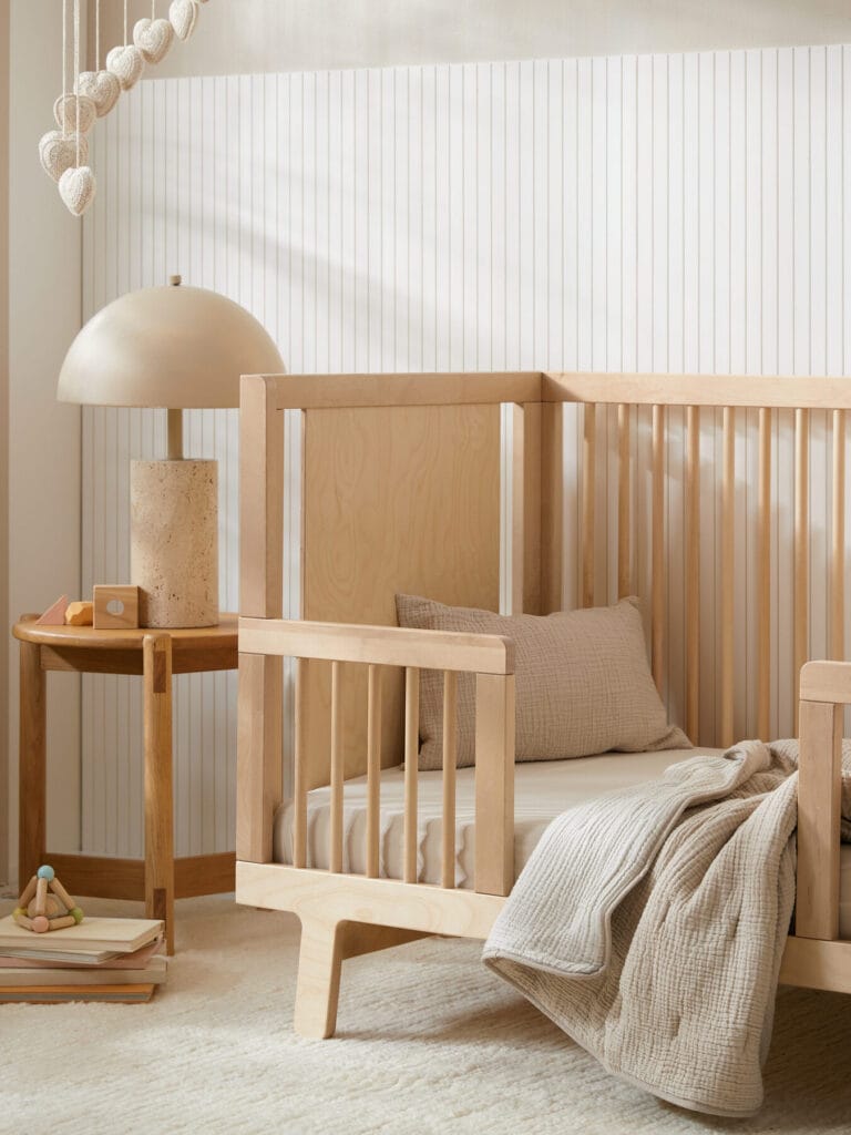 A modern nursery with a light wood crib, beige bedding, a wooden side table with a lamp, toys, and neutral decor on a textured rug.