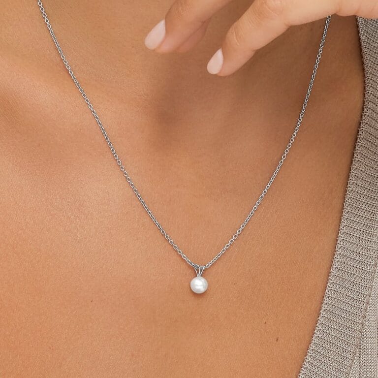 A close-up of a person wearing a silver chain necklace with a single round pearl pendant, shown against bare skin and part of a beige top.