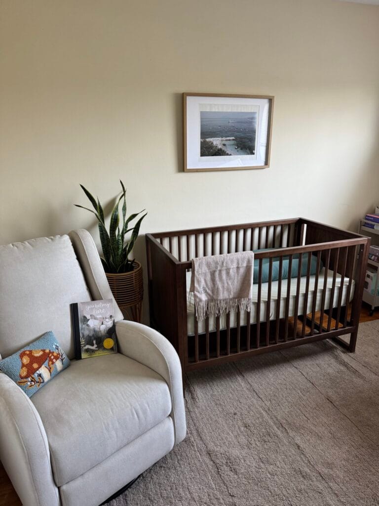A nursery with a dark wood crib, a beige armchair with pillows and a book, a potted snake plant, a framed photo on the wall, and a light rug on the floor.