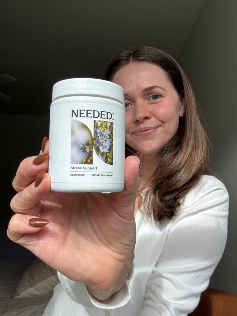 A woman in a white shirt holds up a bottle labeled “Needed: Stress Support” dietary supplement, smiling at the camera.