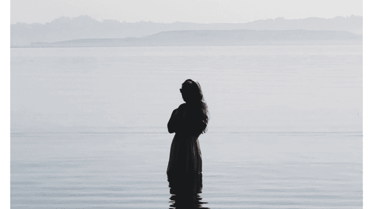 Woman in a dress standing in a sea that's smooth just before sunset with clouds