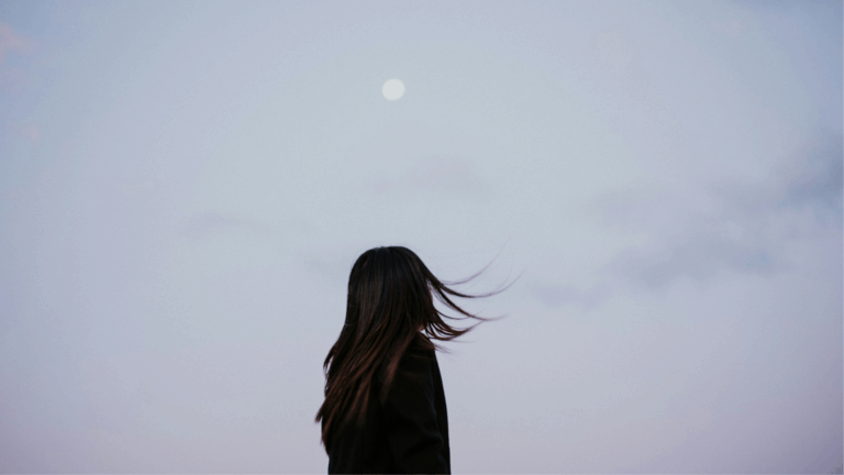 Woman standing outside with her wind blowing in the breeze during a late afternoon with the full Moon in the sky while she thinks about her weekly horoscope