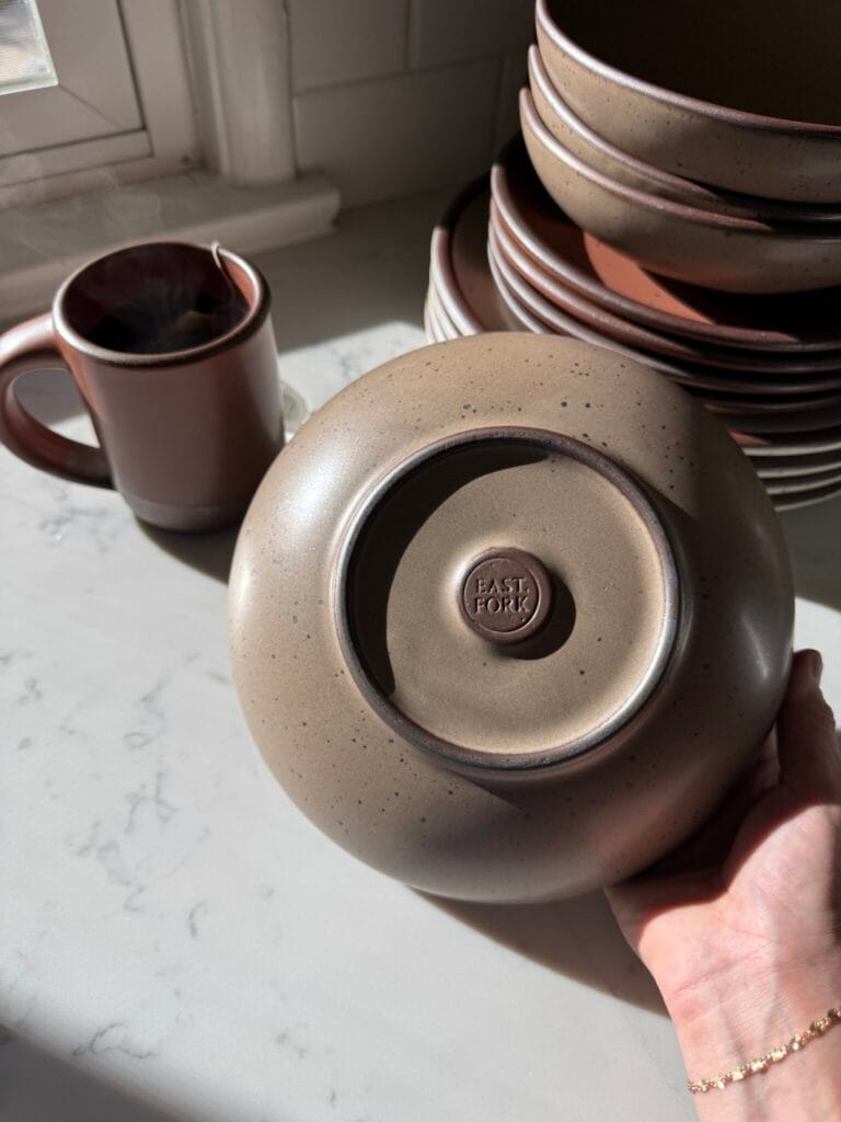 A hand holds an upside-down ceramic bowl with the "East Fork" logo, next to a mug and a stack of plates on a sunlit countertop.