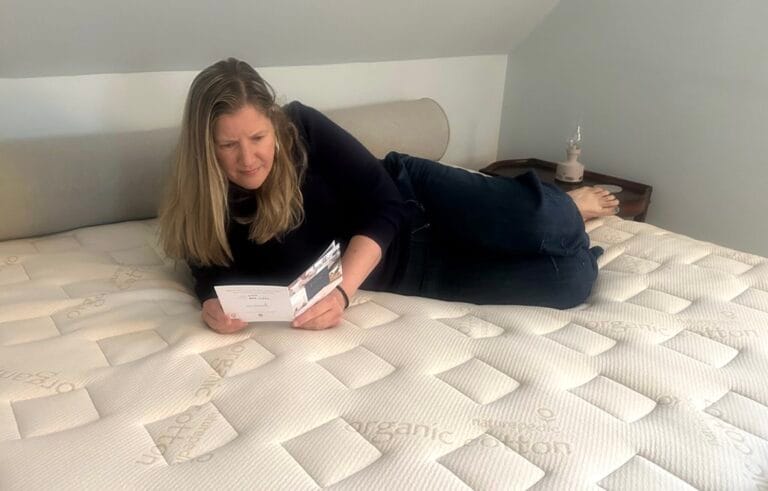 A woman lies on a bed reading a pamphlet. The mattress surface is labeled "organic cotton." A nightstand with a lamp is nearby.