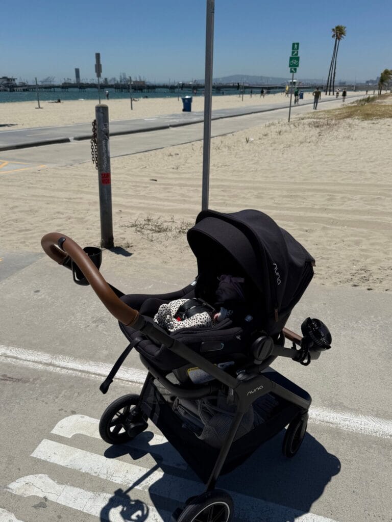A black baby stroller is parked on a paved path by a sandy beach under a clear blue sky.