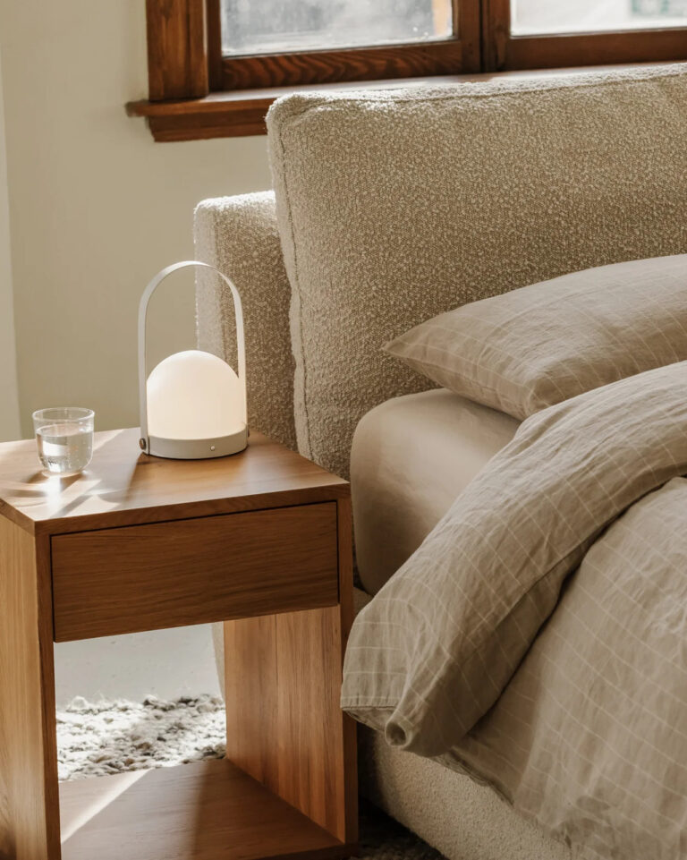 A wooden bedside table with a glass of water and a modern lamp beside a bed with beige linens in a softly lit room.