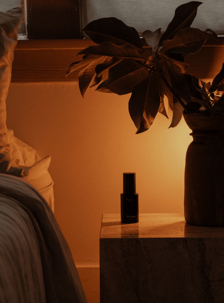A small bedside table with a dark spray bottle and a vase holding leafy branches, lit by warm ambient light next to a bed.