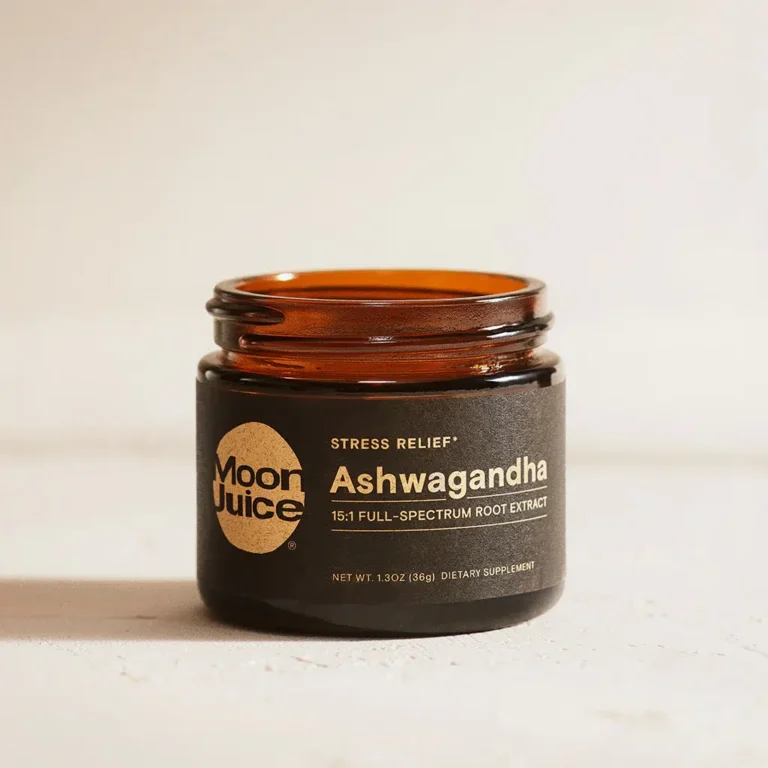 A brown glass jar labeled "Moon Juice Ashwagandha 15:1 Full-Spectrum Root Extract" sits on a light surface. The label notes it is a dietary supplement for stress relief.