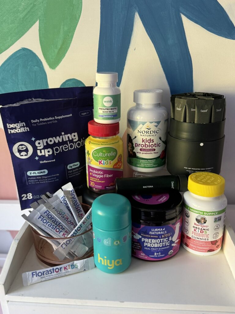 A variety of kids’ probiotic and prebiotic supplements, including powders, chewables, and gummies, are displayed on a white shelf in front of a colorful painted wall.