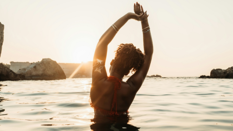 Woman with curly hair pulled back with a tie while she's in the sea and stretching her arms upward