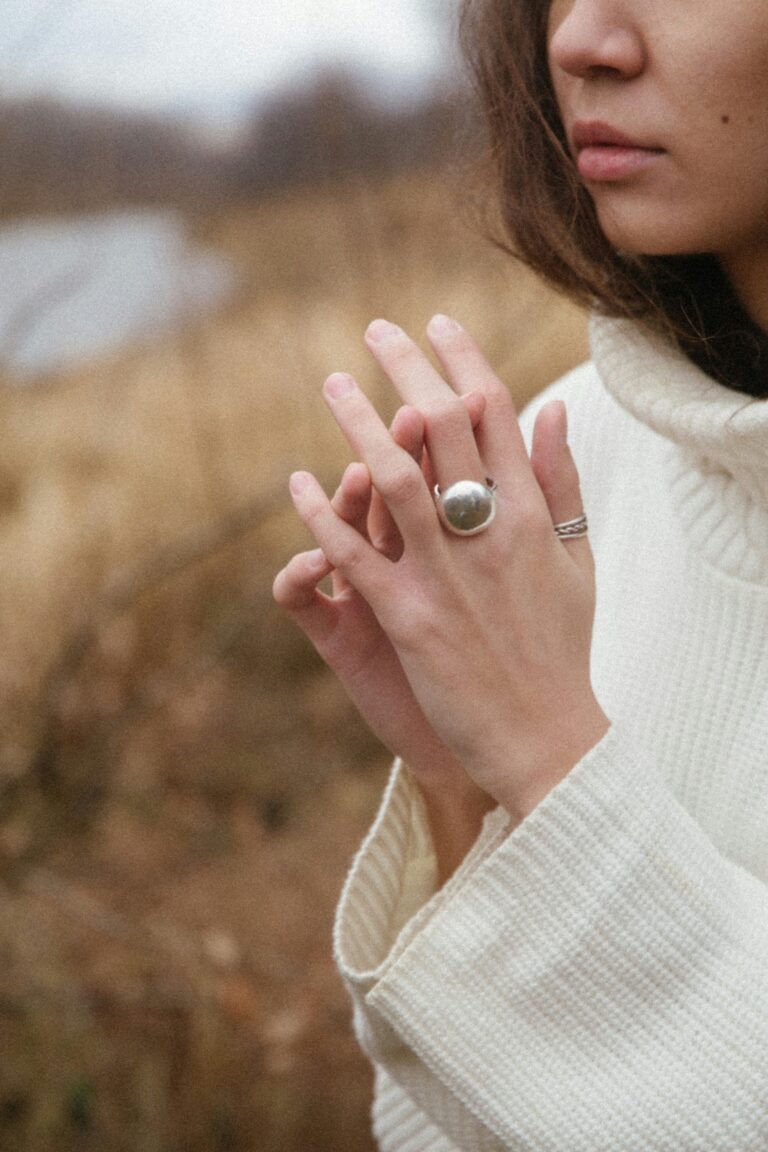 How To Exercise Your Problem-Solving Muscles A person in a cream sweater outdoors touches their fingers, wearing a large silver ring and a thin band ring, with a blurred natural background.