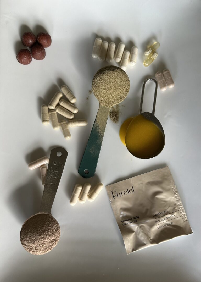 Assorted pills, capsules, powder-filled measuring spoons, and a Perel packet are arranged on a white surface.
