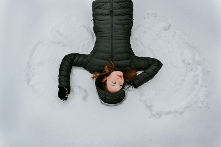 Woman in black winter coat making snow angels in snow.
