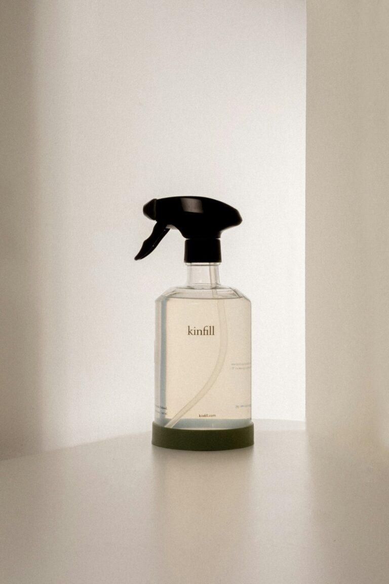 A clear spray bottle labeled "kinfill" with a black nozzle, placed on a white surface against a softly lit background.