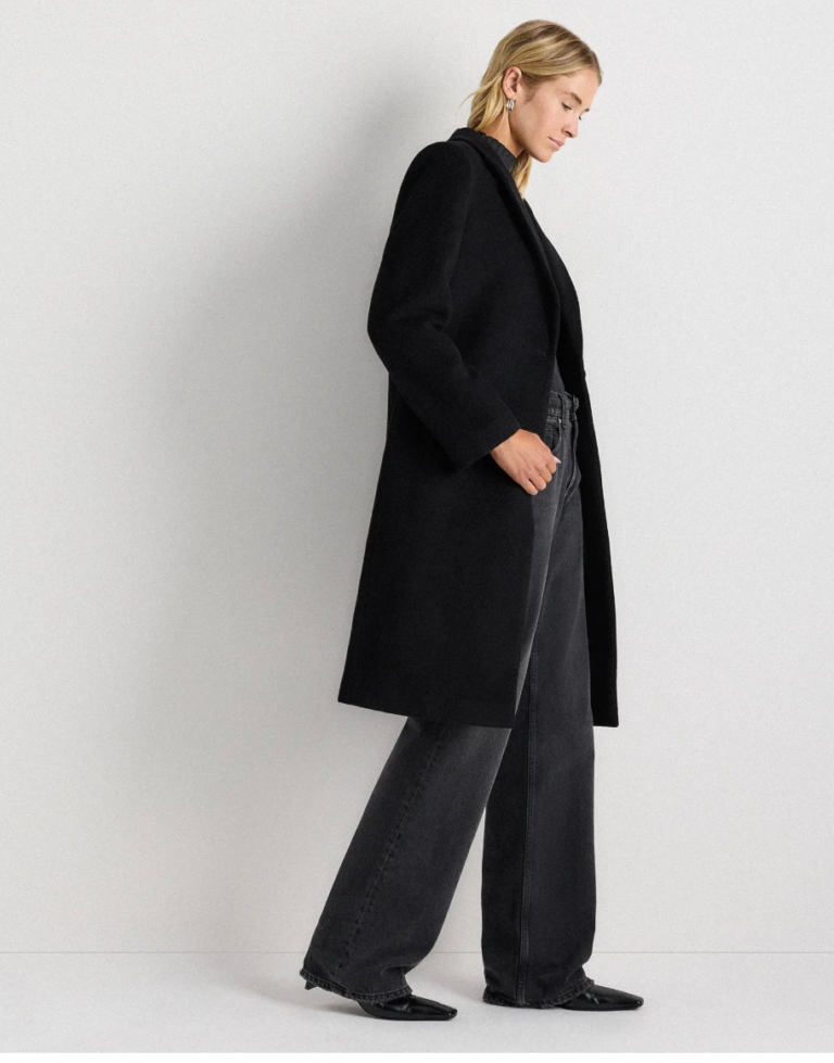 A person with light hair wears a long black coat, dark wide-leg jeans, and black pointed shoes, standing against a plain white wall.