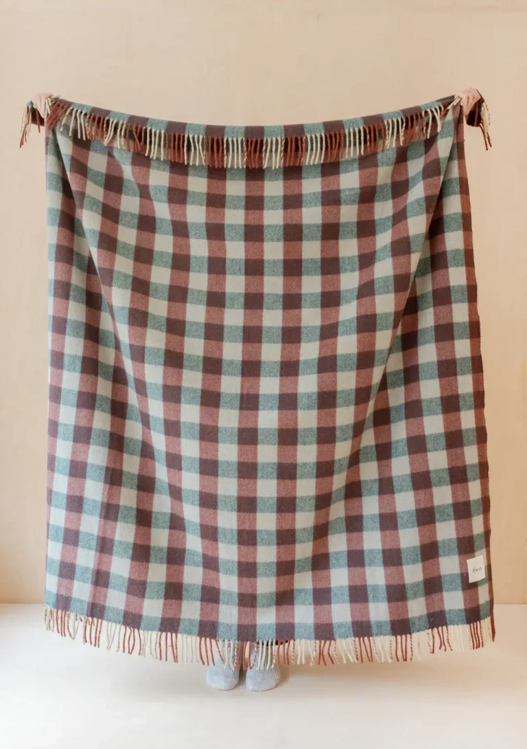 A person stands behind a large, fringed checkered blanket in muted blue and brown tones, with only their feet visible below the blanket's edge.