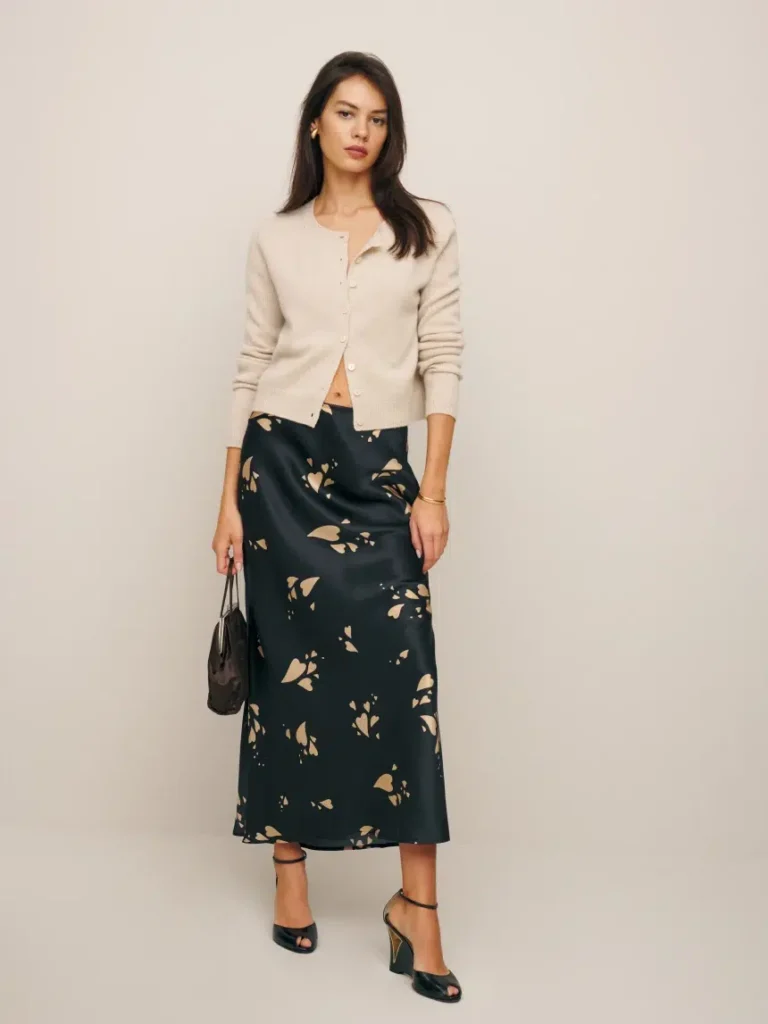 A woman stands against a plain background, wearing a beige cardigan, a black skirt with a beige pattern, and black heels. She holds a small black bag in her left hand.