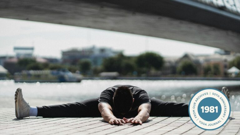 Image of man practicing Wide Legged Seated Forward Fold with a bridge and water in the background.