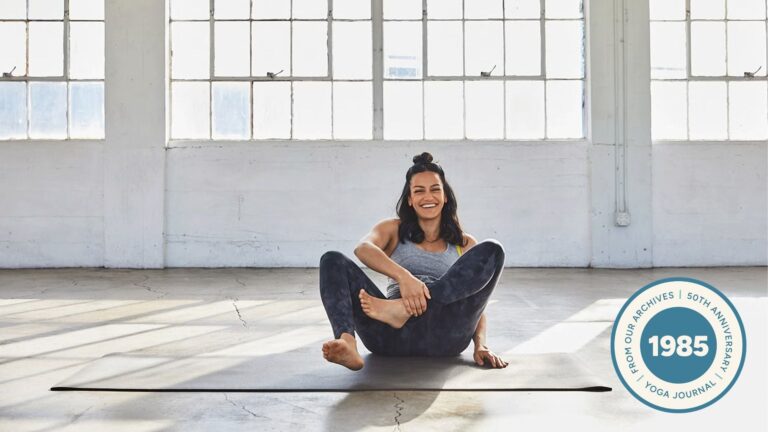 Woman sitting and laughing on a yoga mat as she tries to come into Lotus Pose