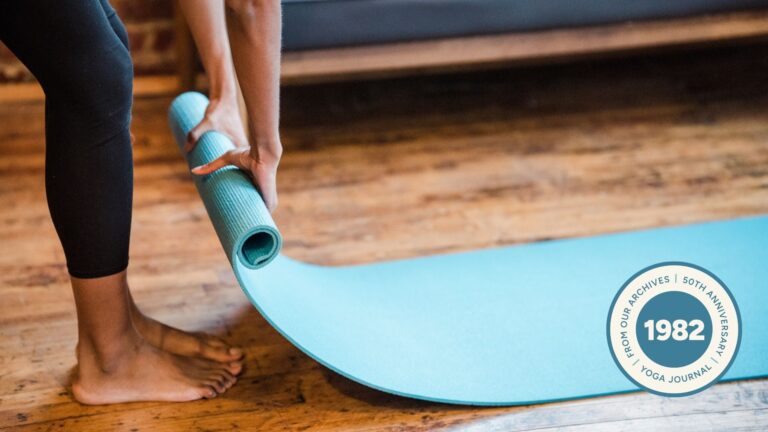 Woman rolling out a blue yoga mat onto a wooden floor.
