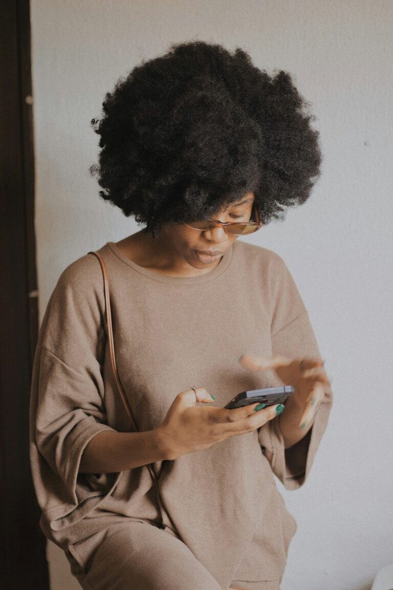 A person with curly hair and glasses is wearing a loose, beige outfit and using a smartphone while standing indoors against a plain, light-colored wall.