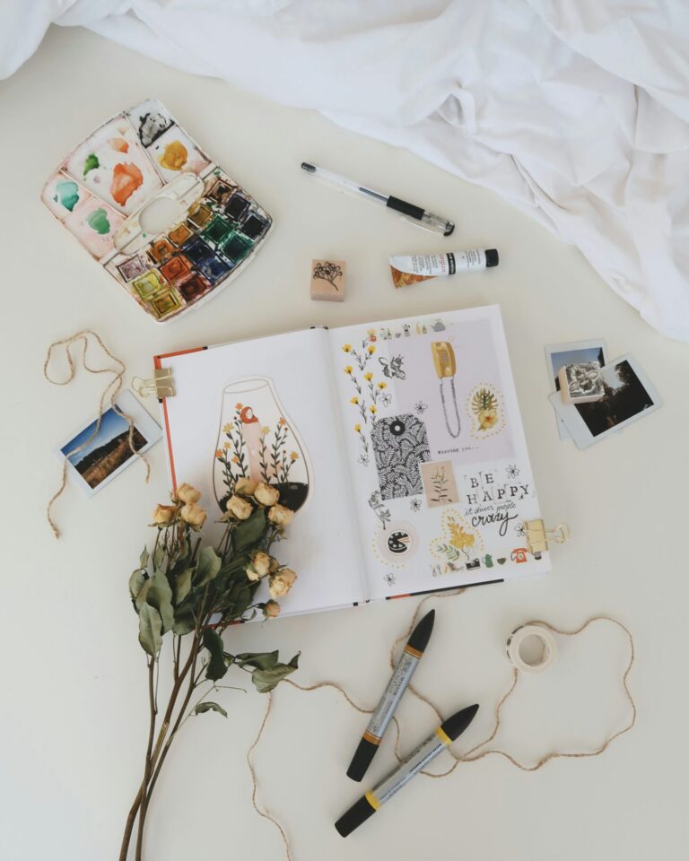 An open sketchbook with drawings and stickers lies on a desk surrounded by markers, dried flowers, watercolor paints, photos, string, and art supplies.