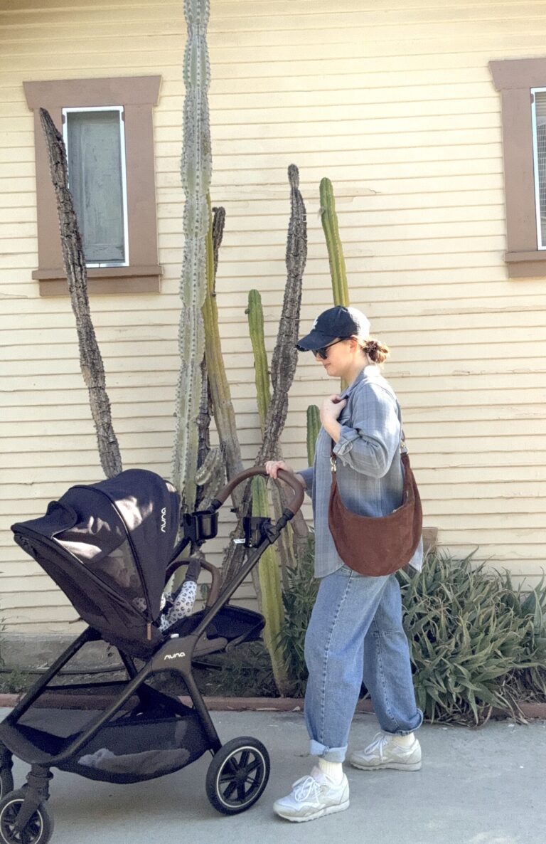 A woman wearing jeans, a cap, and sunglasses pushes a stroller with a baby in front of a beige house with cacti and two windows.