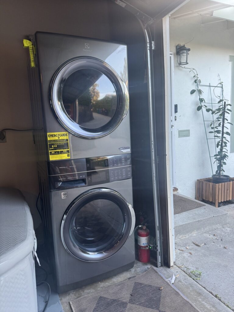 A stacked washer and dryer unit in a garage next to a fire extinguisher, with a house entrance and potted plant visible outside.