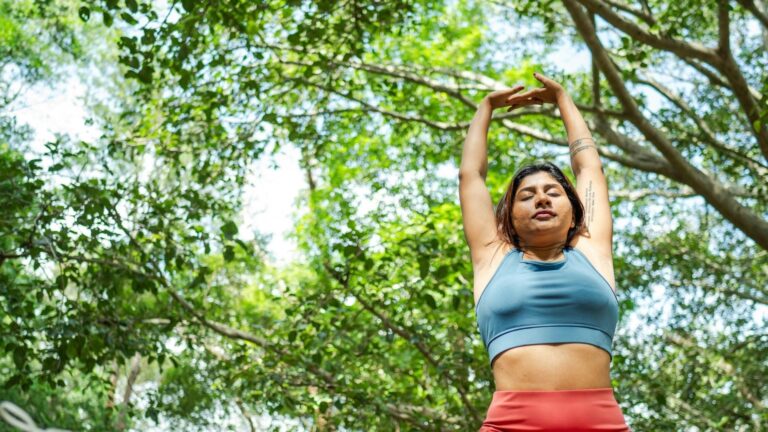 Image of woman in blue sports top and red yoga pants standing outside near green trees reaching her arms up above her head.
