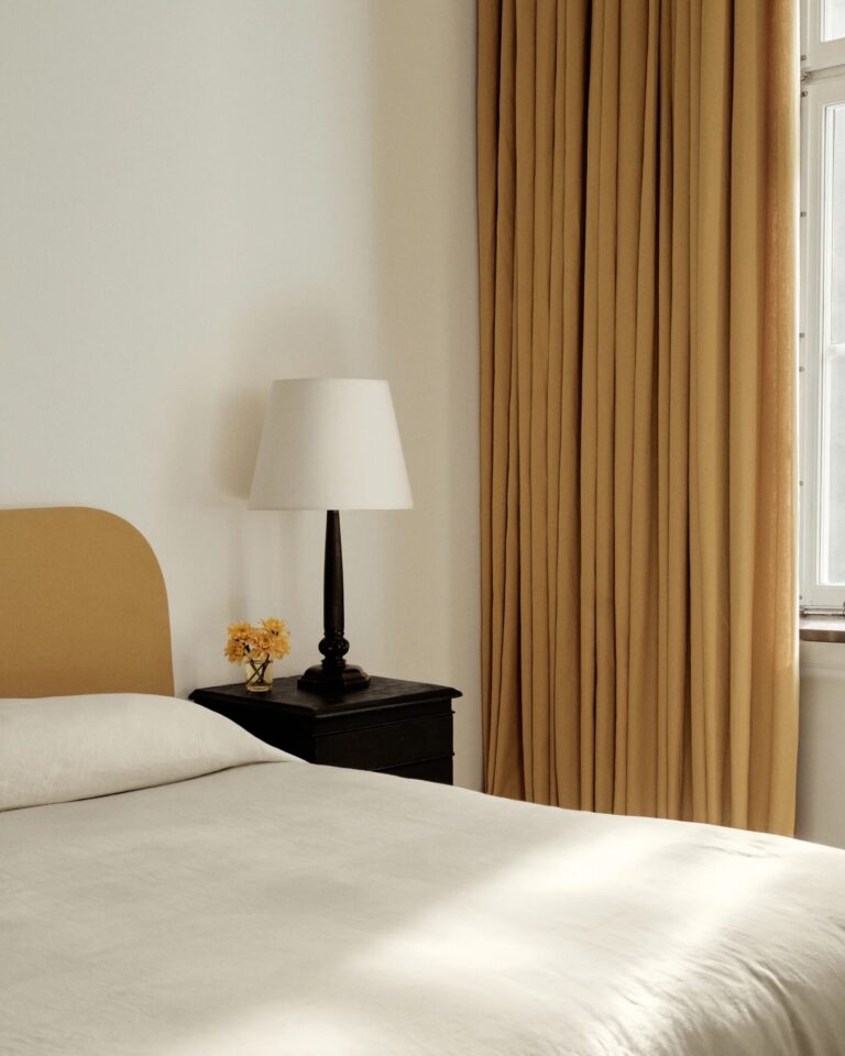 A neatly made bed with a beige headboard, a black nightstand with a lamp and small yellow flowers, and beige curtains beside a window letting in natural light.