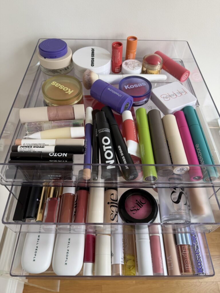 A clear acrylic organizer filled with various makeup products, including lipsticks, powders, mascaras, and compacts arranged neatly in three drawers.