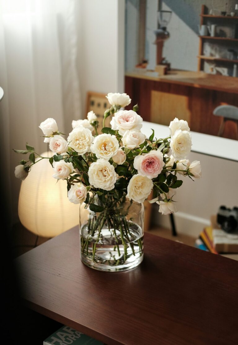 A clear glass vase with white and pale pink roses sits on a wooden table in a softly lit, modern room with a framed photo and lamp in the background.