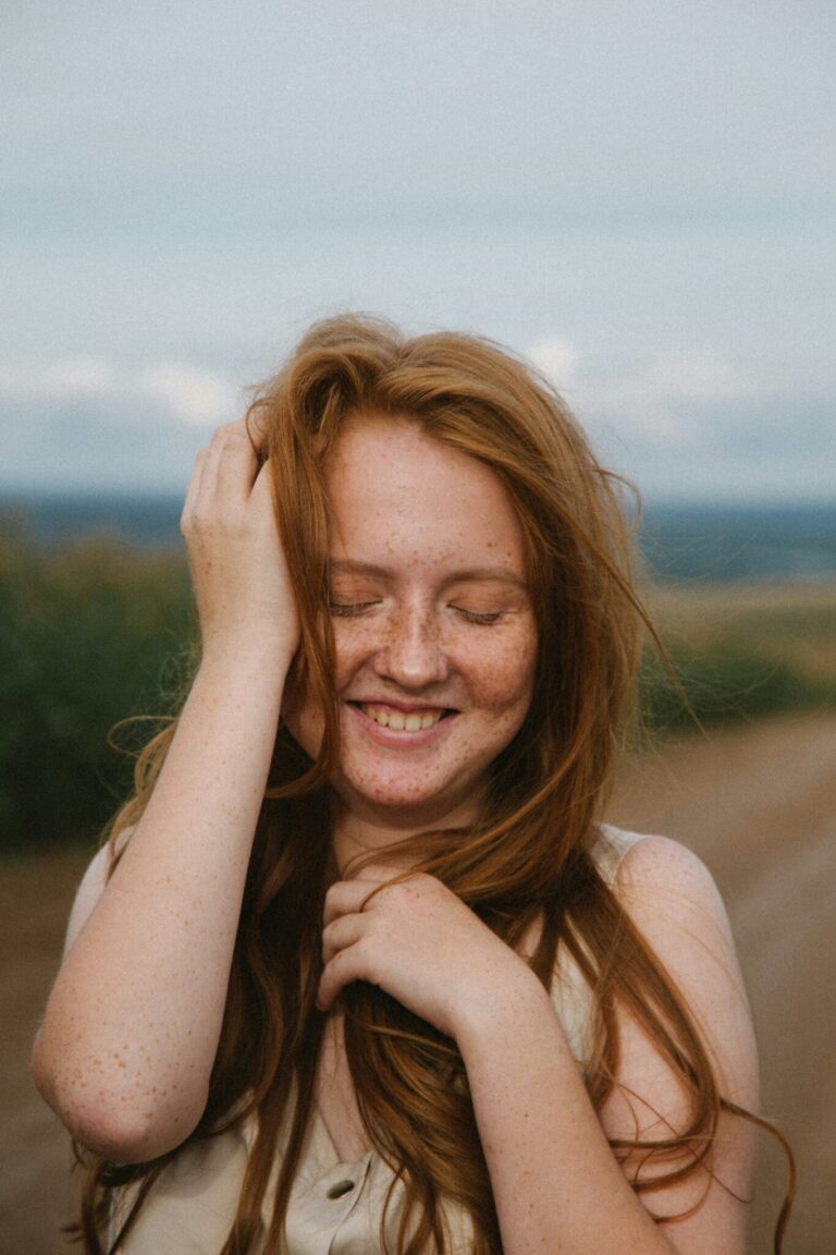 A person with long red hair and freckles stands outdoors on a dirt road, smiling with eyes closed and one hand touching their hair.