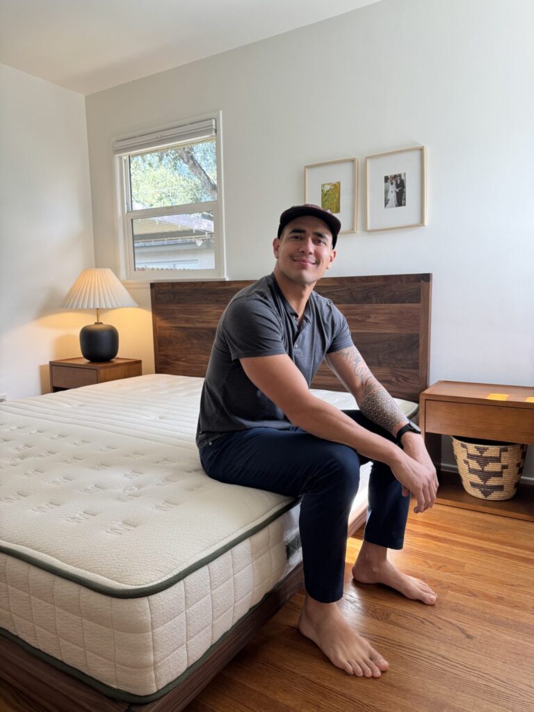 A man sits barefoot on the edge of a bed in a tidy, sunlit bedroom with wooden furniture and framed pictures on the wall.