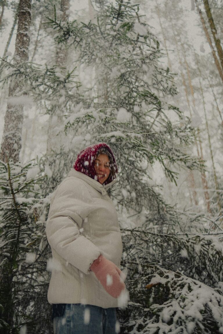 A person in a white winter coat and red hat stands in a snowy forest as large snowflakes fall around them.