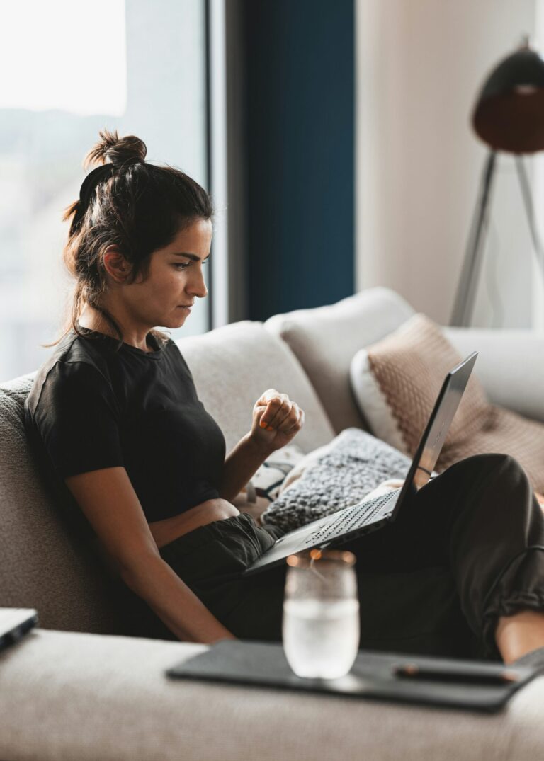 Woman sitting on a couch with a laptop on her lap, focusing on the screen. A glass of water is on the table in the foreground.