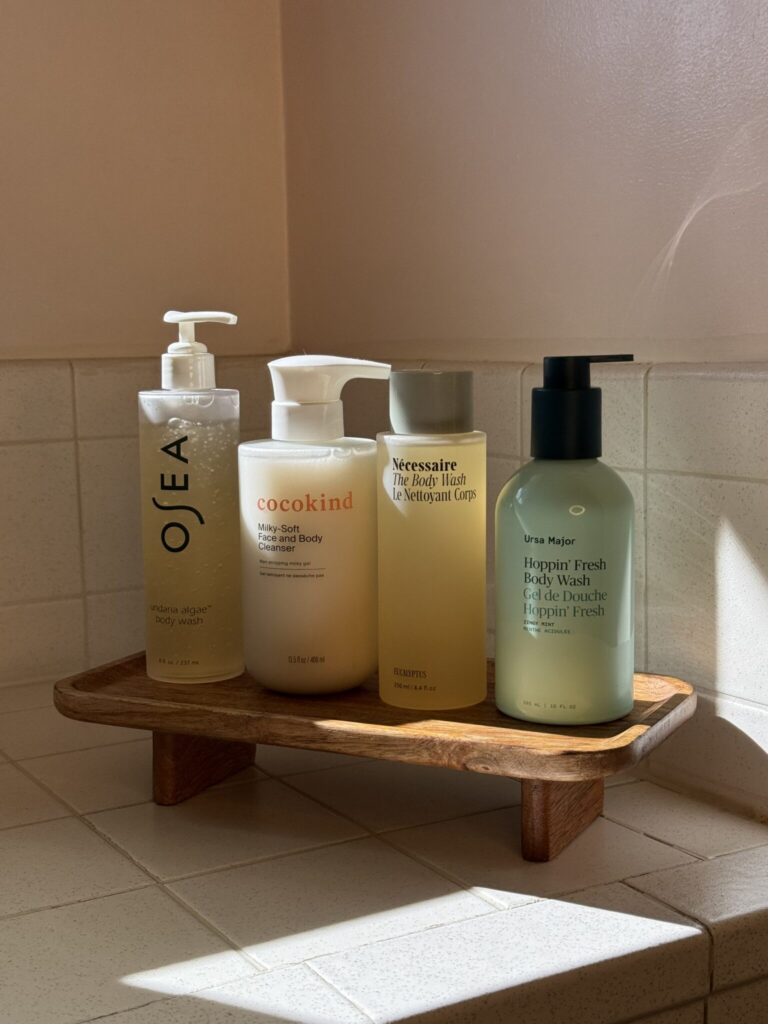 10 Best Natural And Organic Body Washes (2026 Review) Four bottles of body wash and lotion are arranged on a wooden shelf in a tiled bathroom corner, lit by natural sunlight.