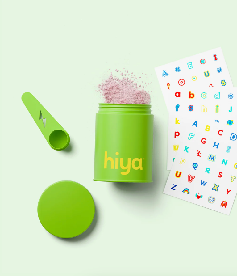 A green container labeled "hiya" with pink powder inside, a matching scoop, a detached lid, and a colorful alphabet sticker sheet on a light green background.