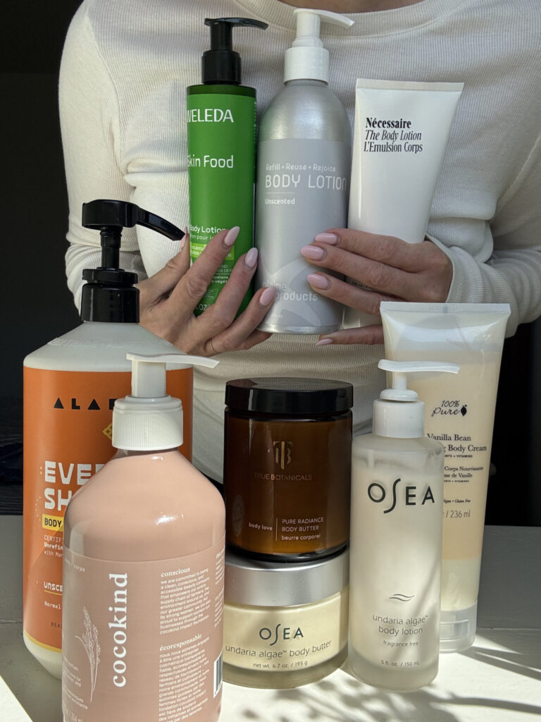 A person holds several bottles of body lotion, with additional skincare and body care products displayed on a table in front of them, all in various shapes and sizes.