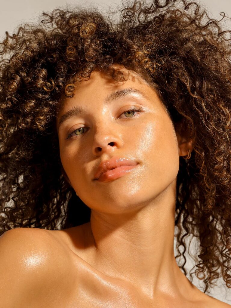 A woman with curly hair and glowing skin looks slightly upward with a neutral expression against a plain background.