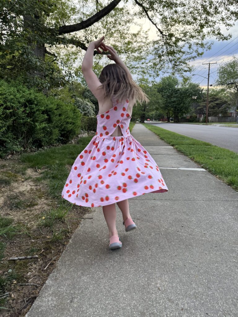 A young girl in a pink dress with red dots spins on a sidewalk near grass and trees on a cloudy day.