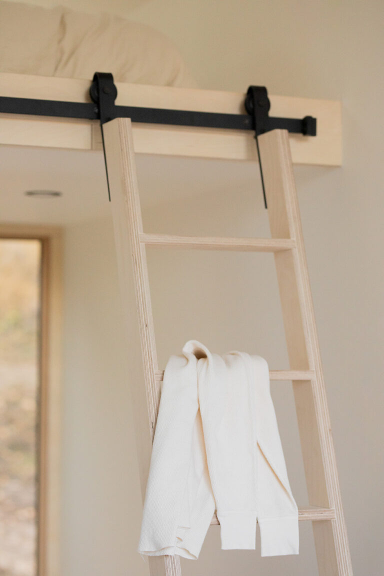 A light wooden ladder leads to a loft bed with bedding. A white sweater is draped over one of the ladder's rungs.