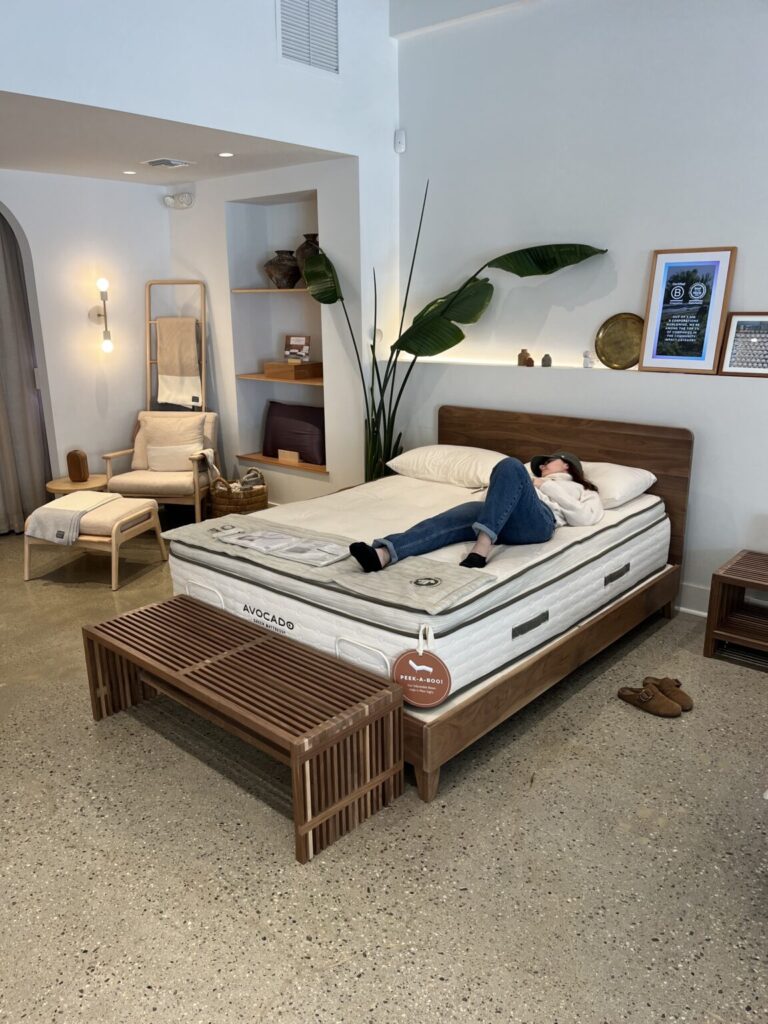 A person lies on a mattress in a modern showroom with wooden furniture, a large plant, framed art on the wall, and a lounge chair in the corner.