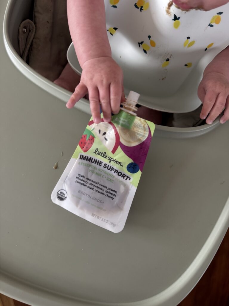 A baby in a high chair holds a Little Spoon Immune Support baby food pouch on a gray tray.