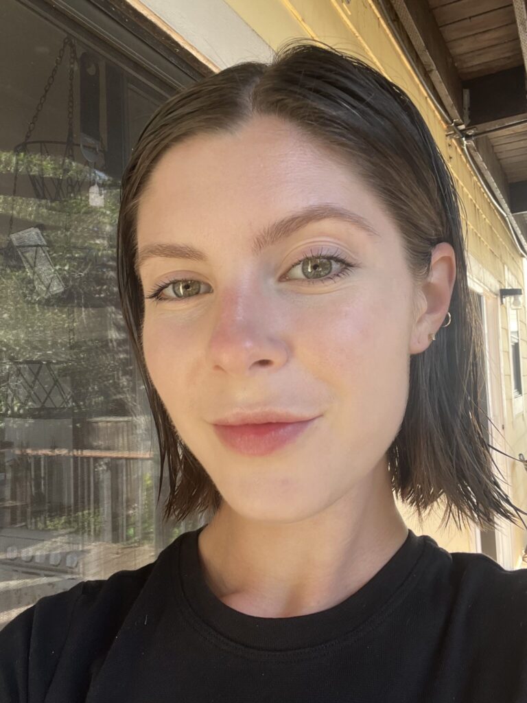 A person with short brown hair and light skin wearing a black shirt stands outside, facing the camera, with a window and yellow siding in the background.