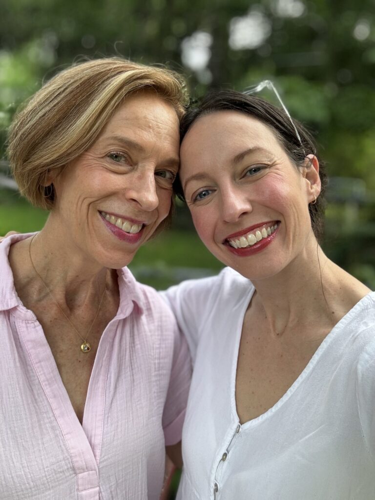 10+ Memorable Mother’s Day Gifts From Sustainable Brands (2026) Two people smiling outdoors, standing close together, wearing light-colored shirts, with greenery in the background.